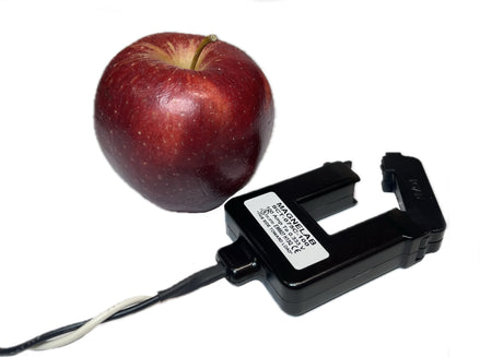 A black magnetic clamp-on current sensor with a white label and attached wire, shown next to a red apple for scale on a white background.